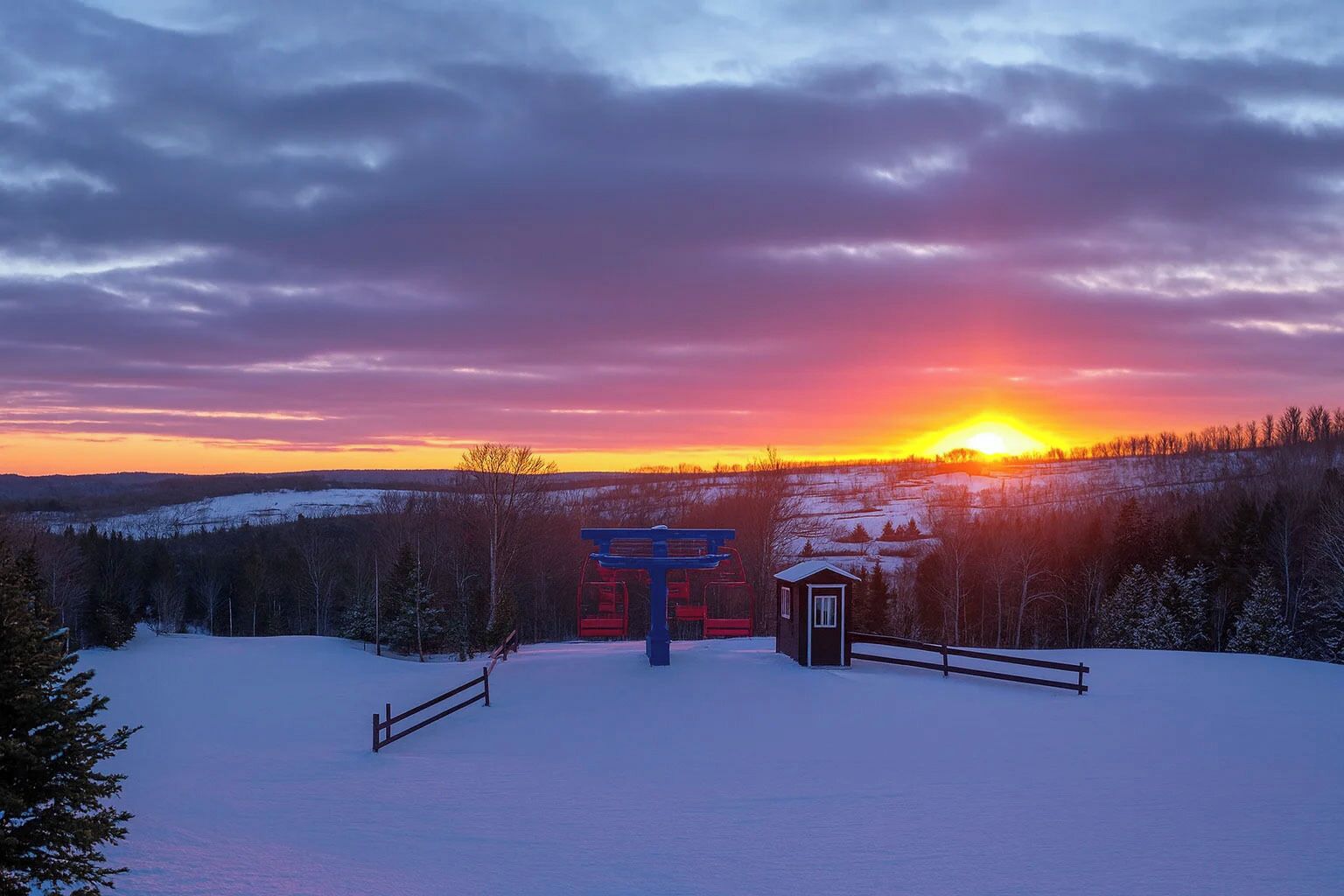 Et sneklædt landskab med en skilift og en lille hytte i solnedgang.