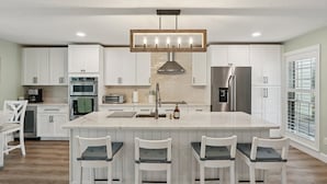 A modern kitchen with white cabinetry, a central island with bar stools, stainless steel appliances, and a pendant light fixture. - Tee to Sea Retreat - Inside TPC Sawgrass Gated Community, Ponte Vedra Beach  (Palm Valley)