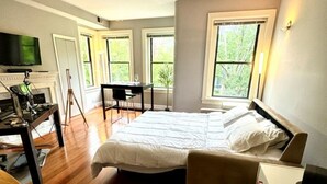 Een slaapkamer met een groot bed, een bureau en een stoel. Er zijn ramen en een televisie aan de muur.