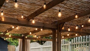 Un patio al aire libre con una pérgola de madera, luces colgantes y un sofá modular gris con cojines.