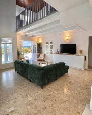 A spacious living area with a green sectional sofa, a flat-screen TV, and a balcony with potted plants. - Magnificent villa with large pool and pleasant living space. (Rivière-Salée)