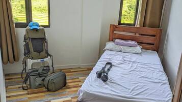 A small, neatly arranged room with a wooden bed, a window with curtains, a backpack, and a camera on the bed.