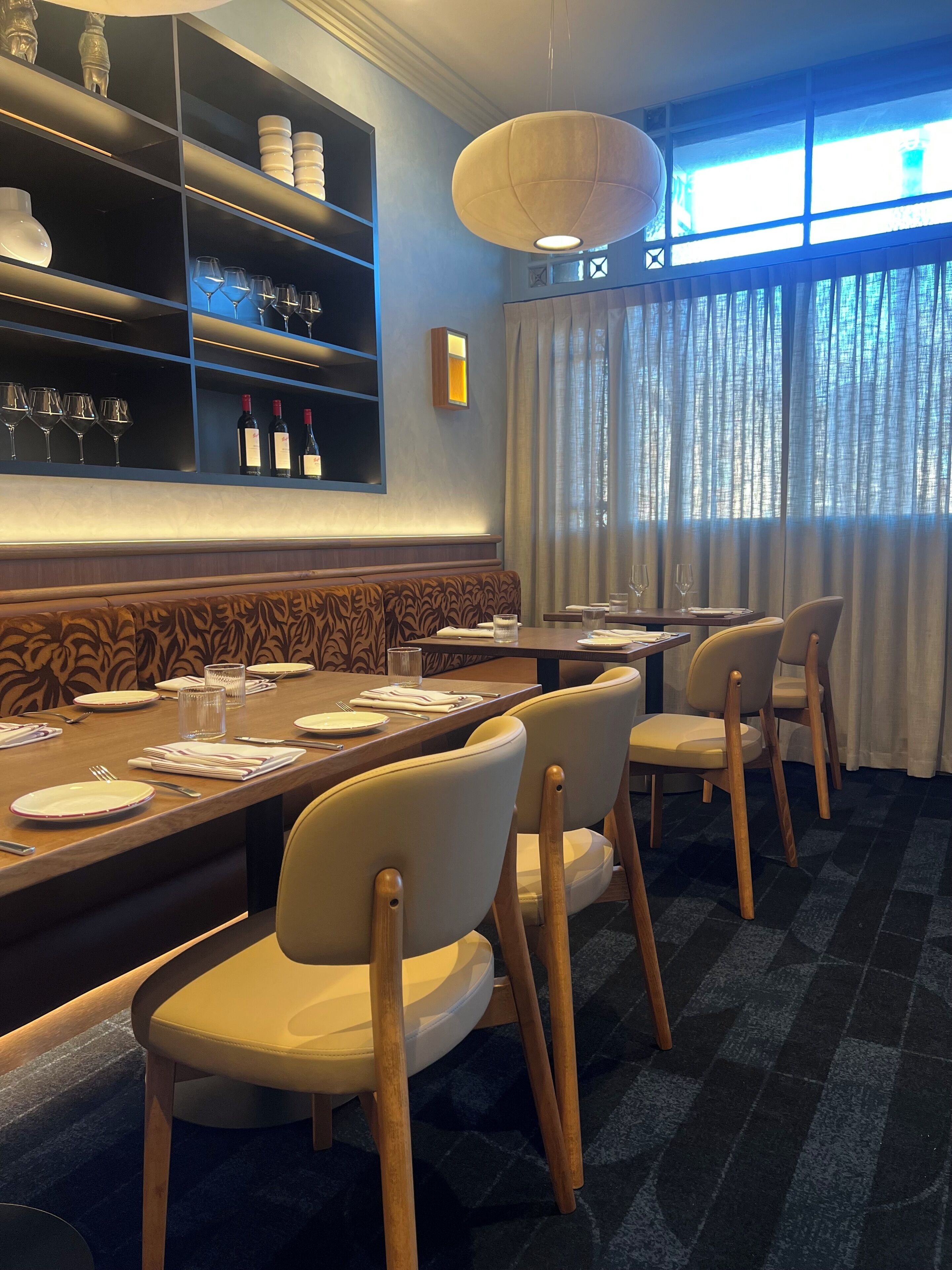 A restaurant interior with a long dining table, chairs, and a bar area with shelves.