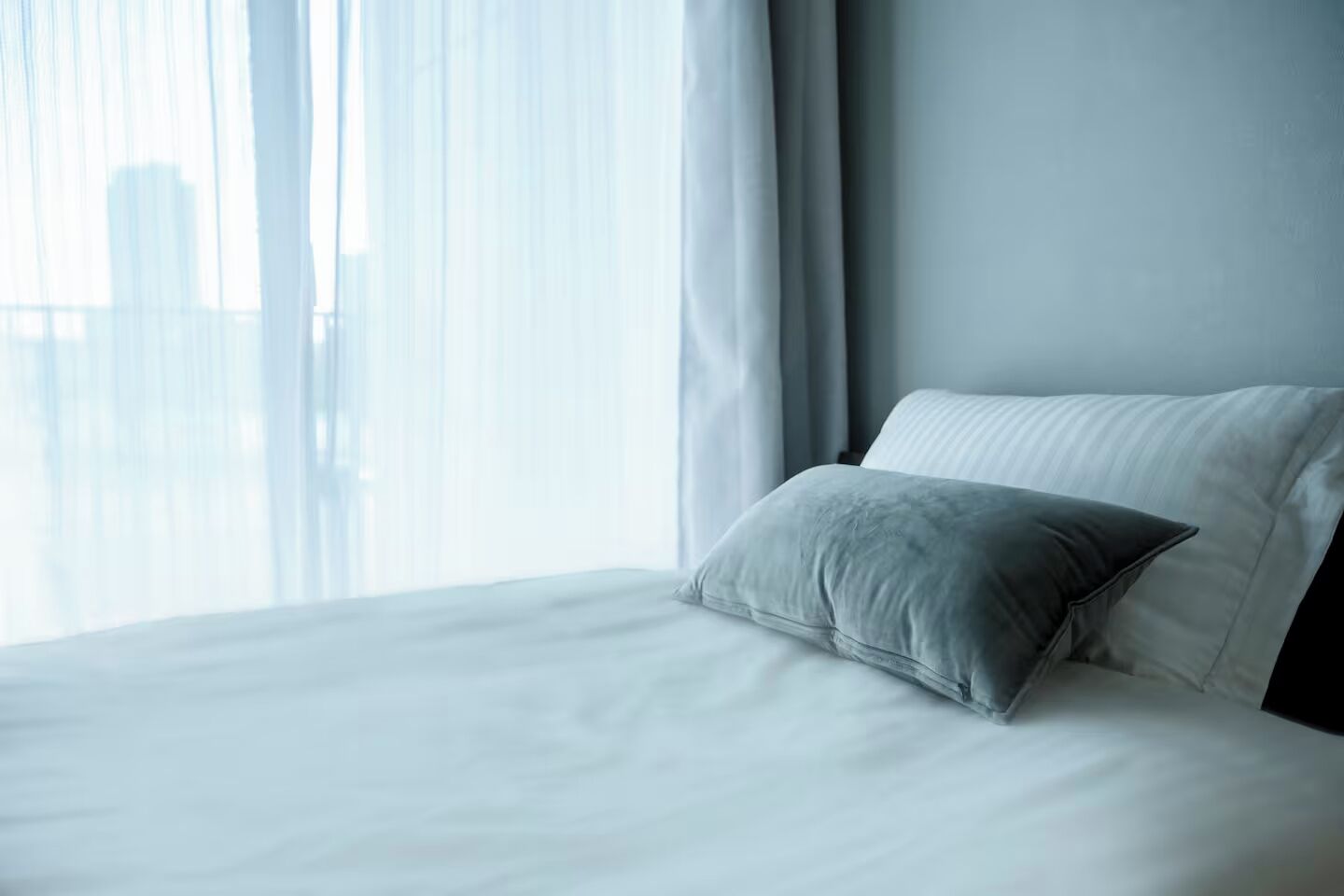 Ein Hotelzimmer mit einem großen Bett, weißen Laken und Blick auf eine Stadtlandschaft durch leichte Vorhänge.