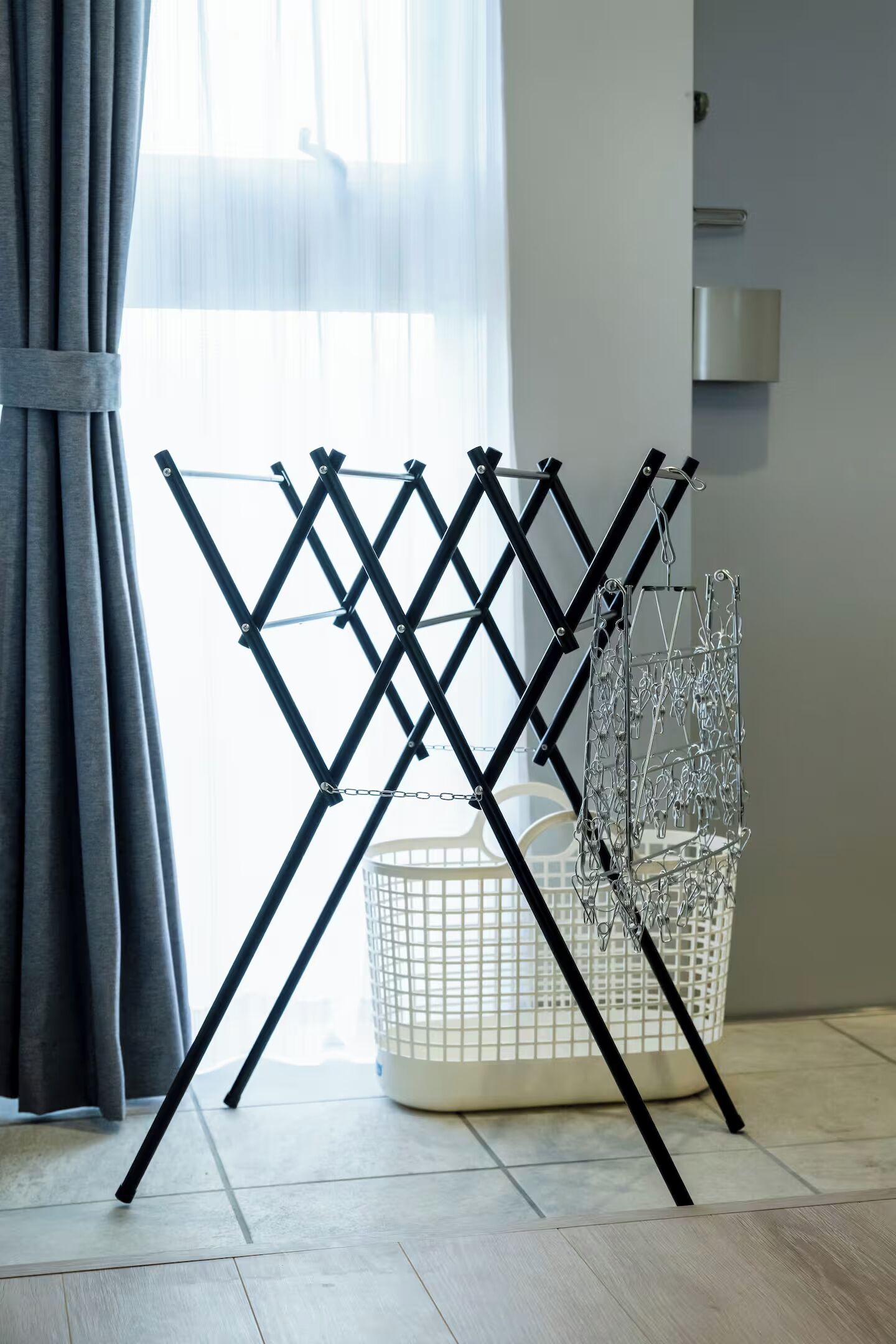 A room with a black drying rack, a white laundry basket, and a window with curtains.