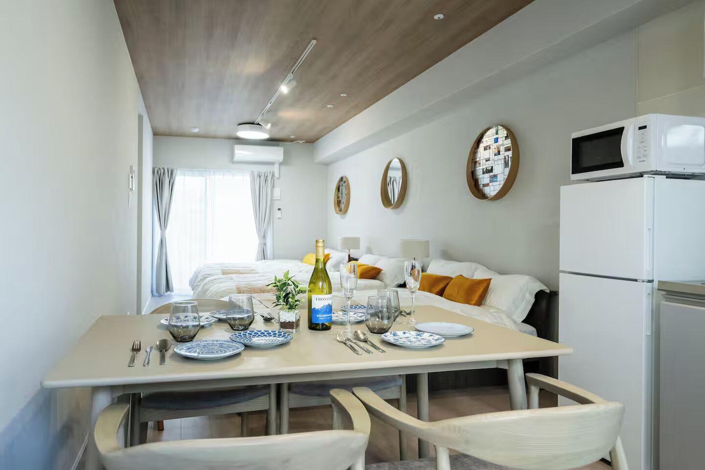 A modern hotel room with a dining area, a bed with white bedding and orange pillows, a white refrigerator, and a white microwave.