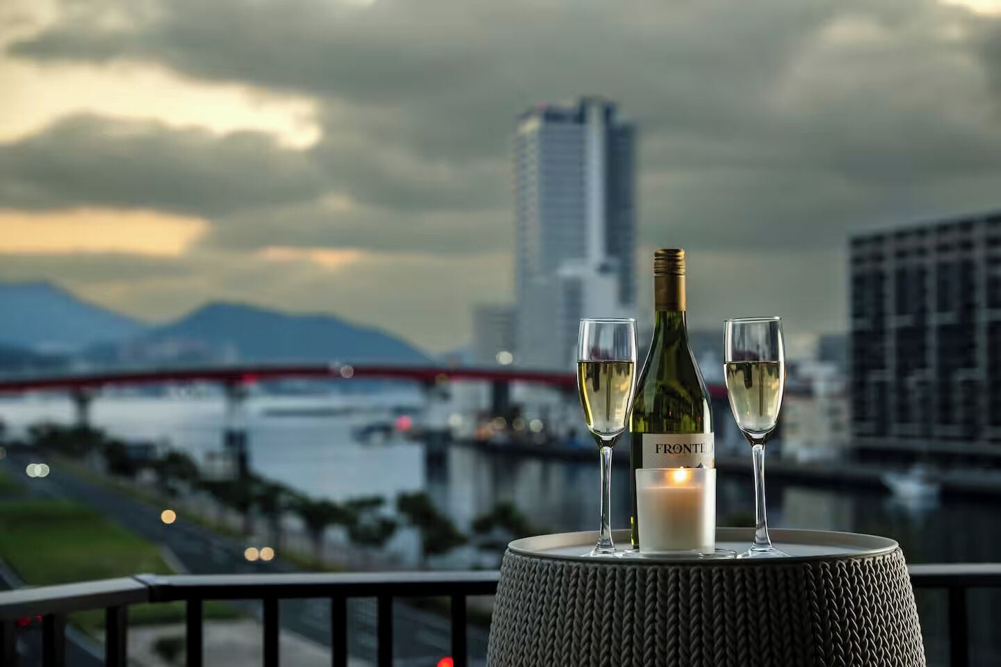 A table with two wine glasses and a bottle on a balcony overlooking a cityscape with a bridge and mountains.