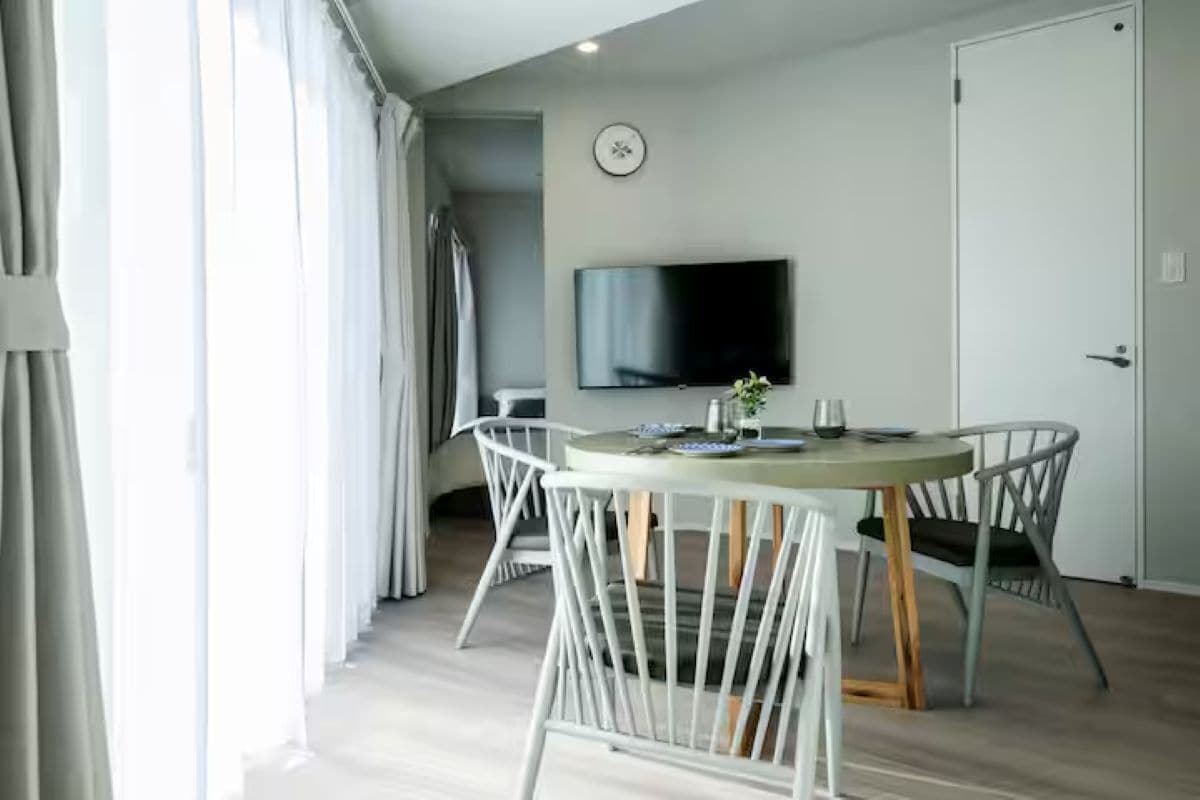 Una habitación moderna con una mesa de comedor redonda, sillas blancas, un televisor de pantalla plana en la pared y una puerta.