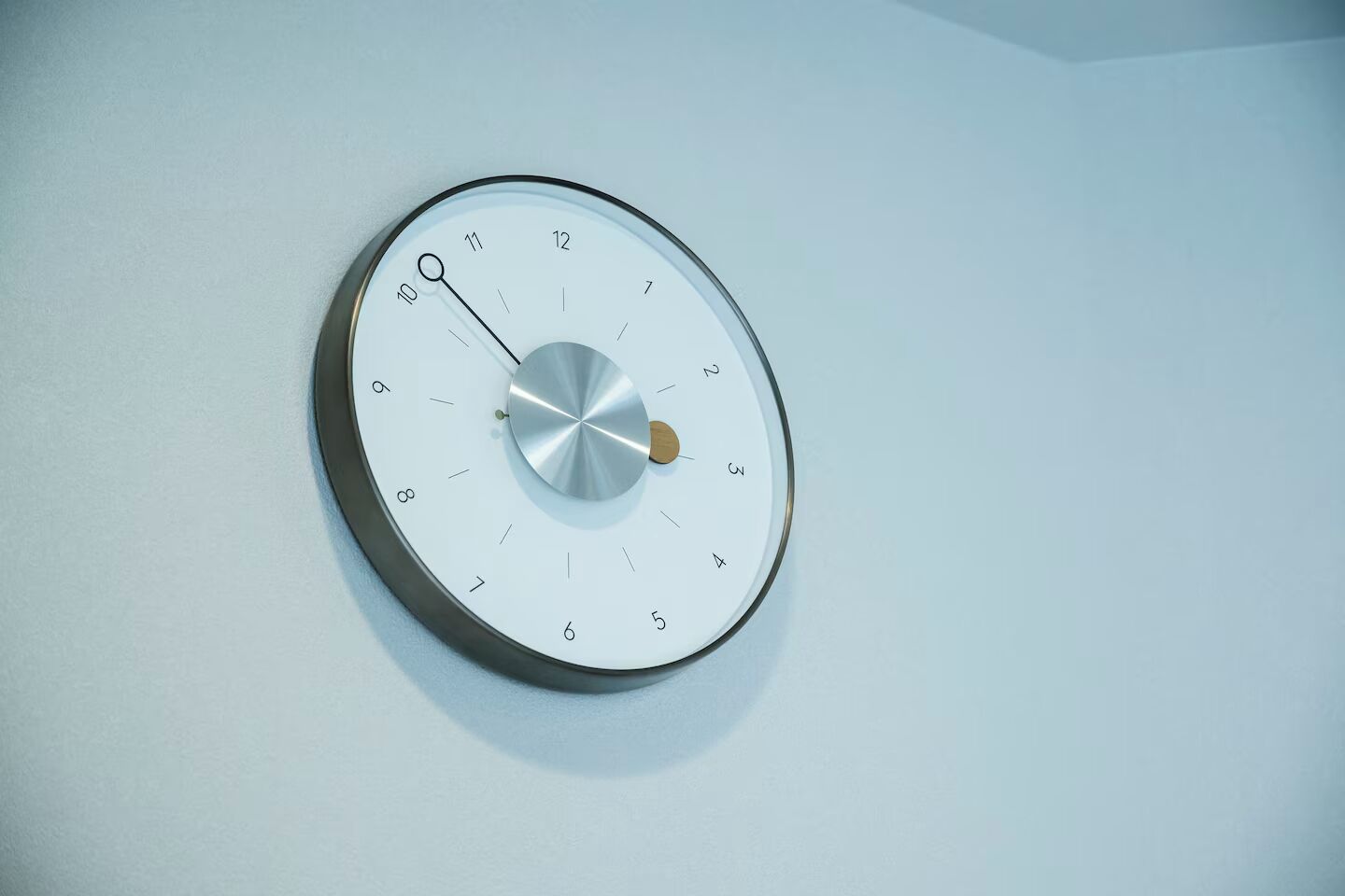 A round wall clock with a minimalist design, featuring a metallic face and a wooden hour hand.