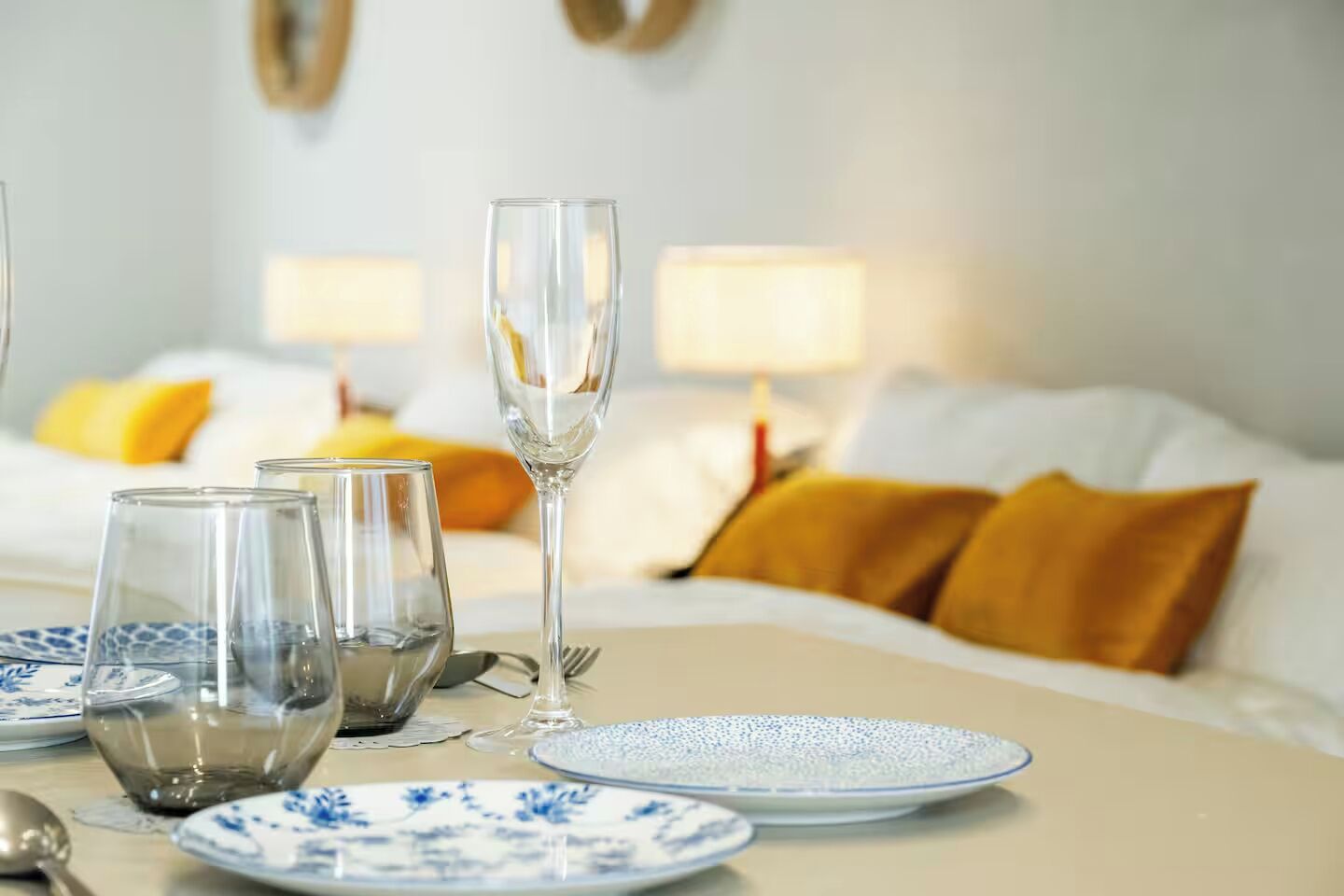 Una mesa de comedor con cristalería y platos transparentes, una cama con almohadillas amarillas y una lámpara al fondo.