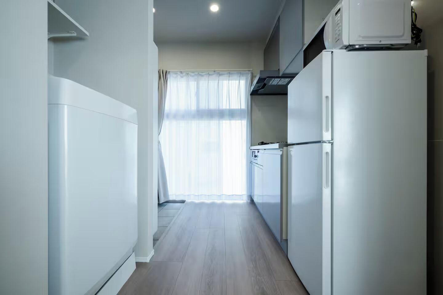 Una cocina estrecha con electrodomésticos blancos, una ventana con cortinas ligeras y suelo de madera.