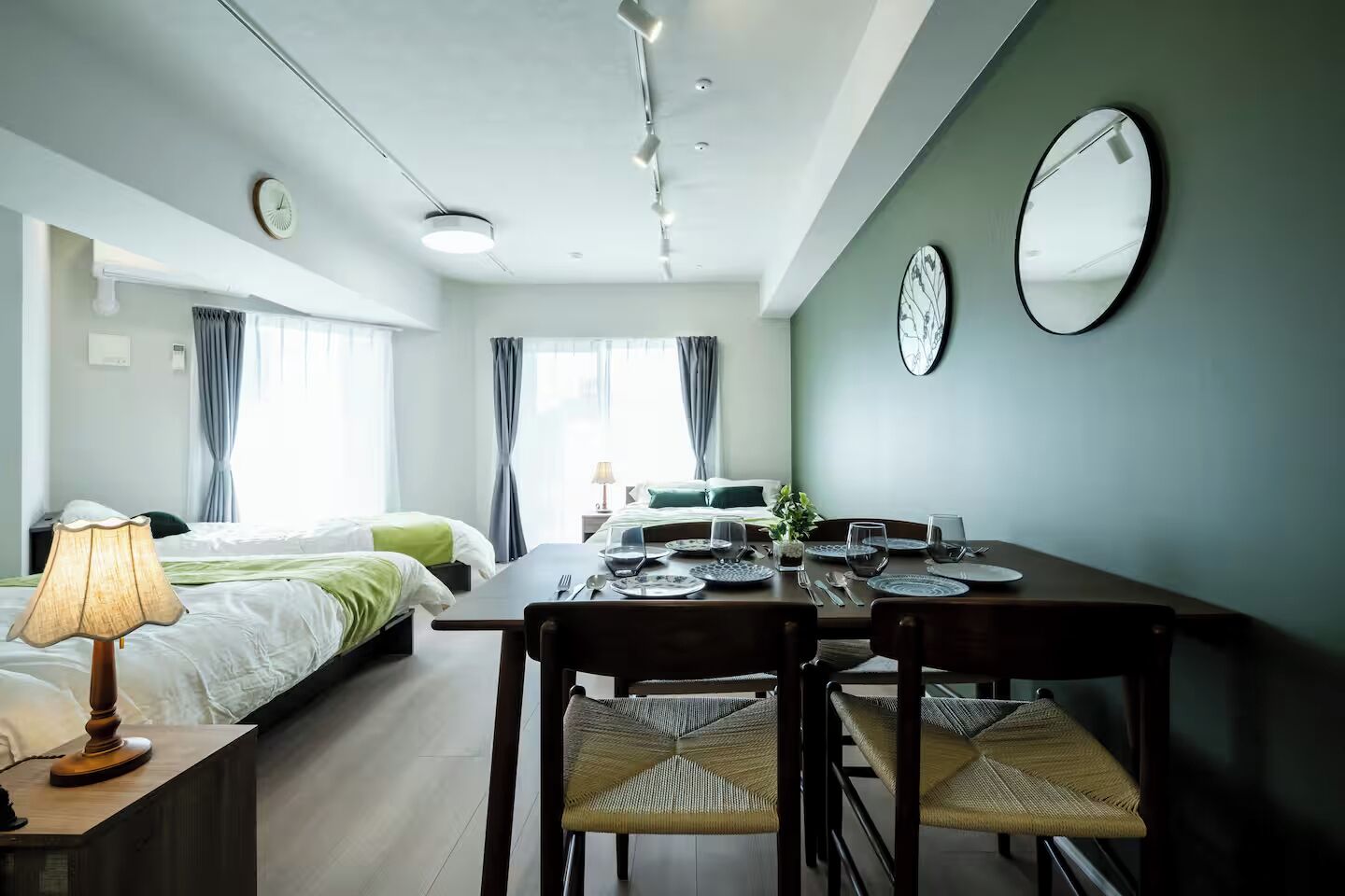 A modern hotel room with a dining area, a bed with green bedding, a bedside table with a lamp, and a large mirror on the wall.
