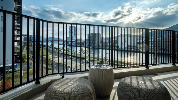 Un balcon offrant une vue sur un paysage urbain, une rivière et des montagnes. On y trouve deux poufs ronds en osier et une table en verre avec deux verres et une plante.