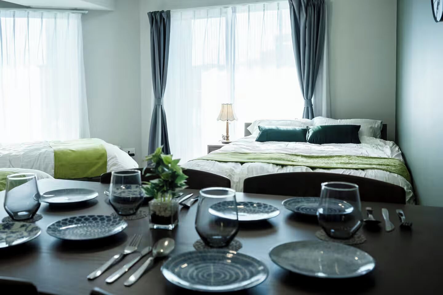 A hotel room with two beds, a dining table set with plates, glasses, and cutlery, and a lamp on a bedside table.