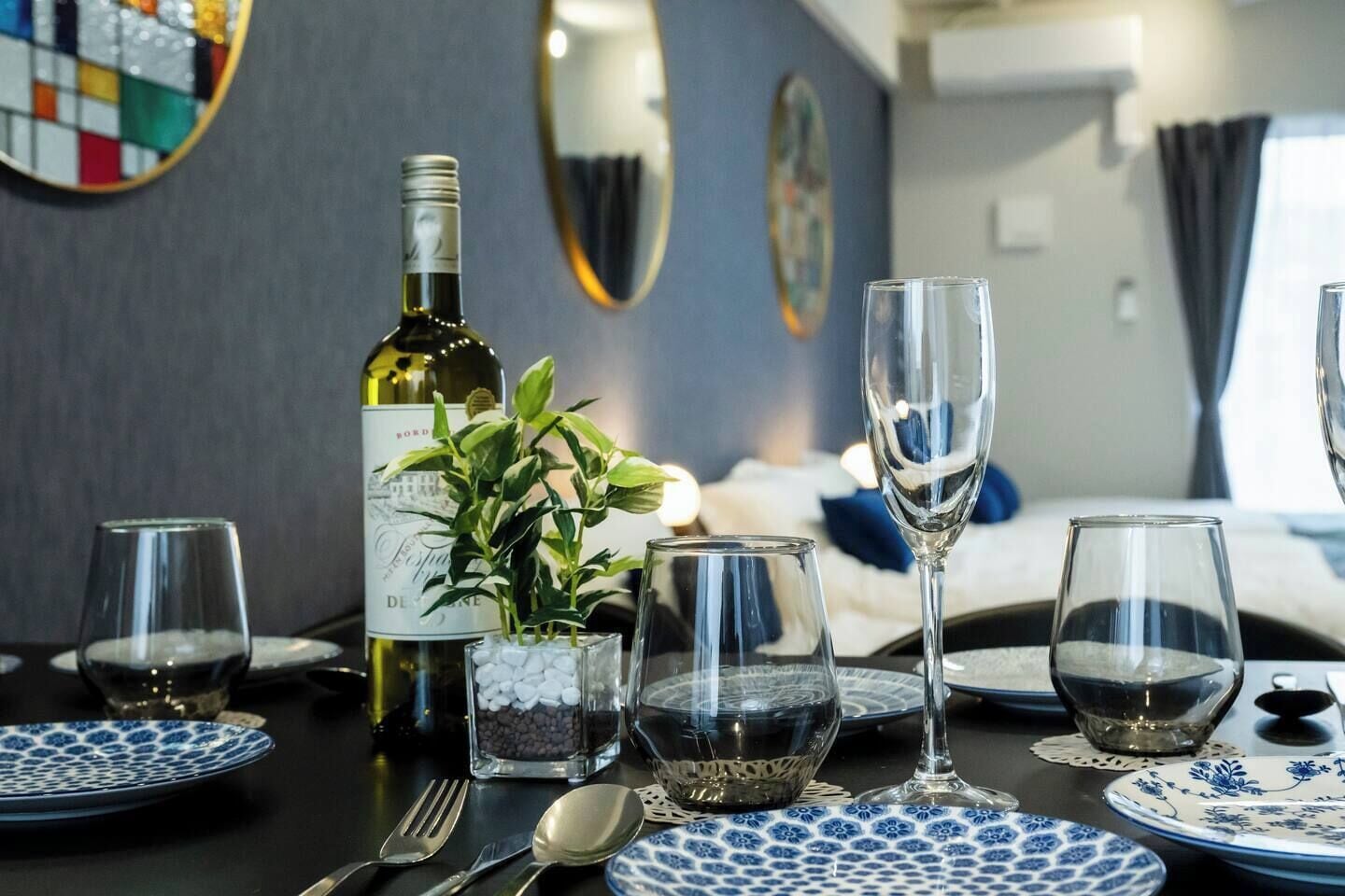 Una mesa de comedor dispuesta con platos, vasos, cubiertos, una botella de vino y una planta decorativa.