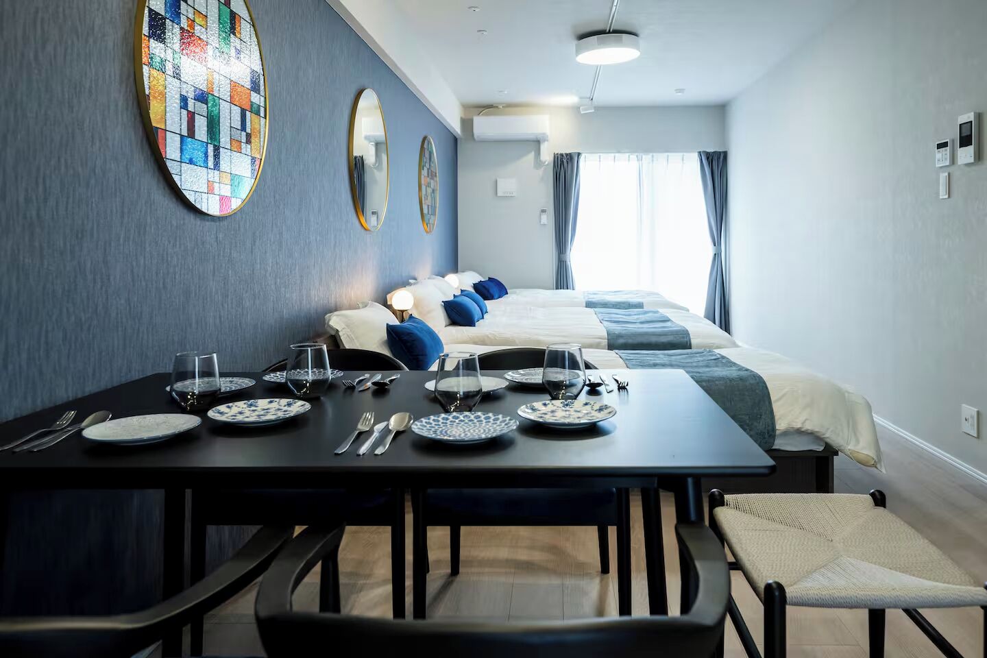 A modern hotel room with a dining area, a bed with blue and white bedding, and a large window with curtains.