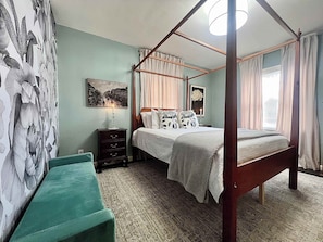 A four-poster bed with a canopy, a nightstand with a lamp, a green bench, and a wall with a floral pattern.