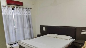 Ένα υπνοδωμάτιο με ένα κρεβάτι, κομοδίνα, μια καρέκλα, ένα κόκκινο κλιματιστικό και ένα παράθυρο με κουρτίνες.