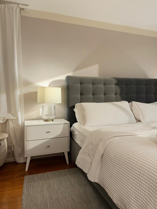 A bedroom with a bed, a nightstand with a lamp, a grey headboard, and a grey rug.