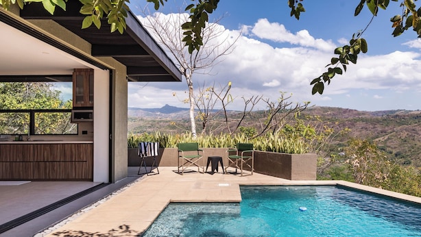 Un área de piscina moderna al aire libre con vistas a montañas distantes, una terraza de madera y una zona de estar con sillas y una mesa.