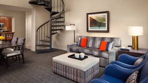 Ein modernes Wohnzimmer mit einem grauen Sofa, einem gestreiften Hocker und blauen Sesseln. Eine Wendeltreppe führt zur oberen Etage.