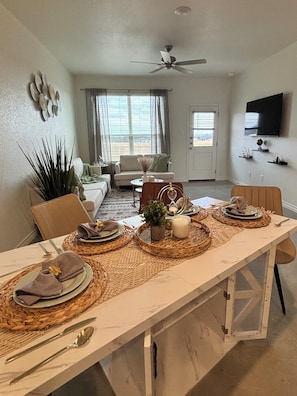 A dining area with a long table set for four, a ceiling fan, and a television mounted on the wall. - 14+ day Stay in Killeen- Fully furnished, all utilities, WiFi, security camera (Killeen)