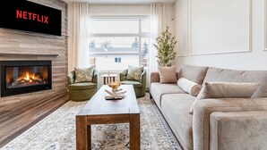 A modern living room with a fireplace, a large flat-screen TV displaying 'NETFLIX', a green armchair, a beige sofa, and a coffee table with books. - Luxury 3BR Retreat Near Downtown | 5-PC King Ensuite +Private Balcony | Sleeps 6 (Calgary)