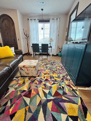 A living room with a patterned rug, a sofa, a coffee table, a dining area with chairs, a cabinet, and a window with curtains. - Songbirds Spot Cottage in a peaceful, upscale area in Layton, UT. (Layton)
