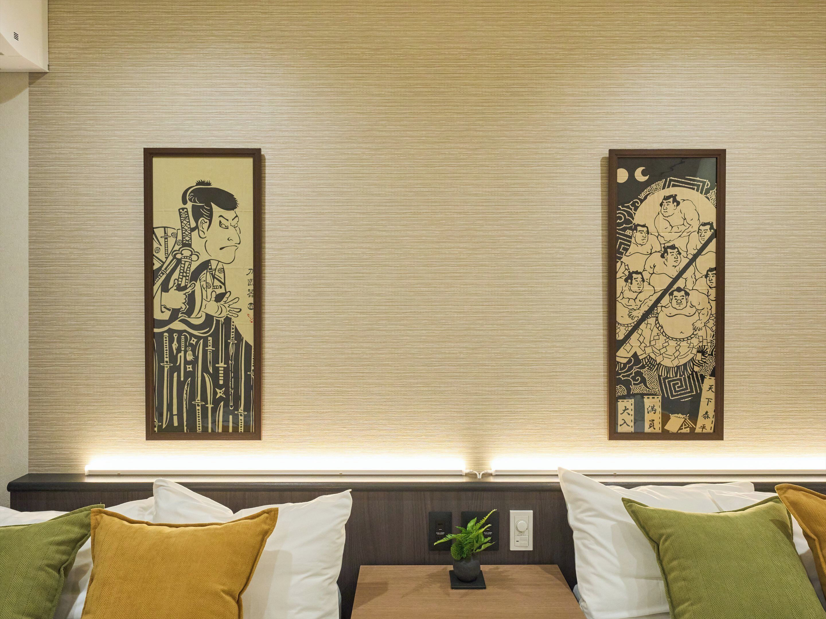 Habitación de hotel con estética japonesa tradicional, que presenta dos obras de arte enmarcadas en la pared y una cama ordenada con varios cojines.
