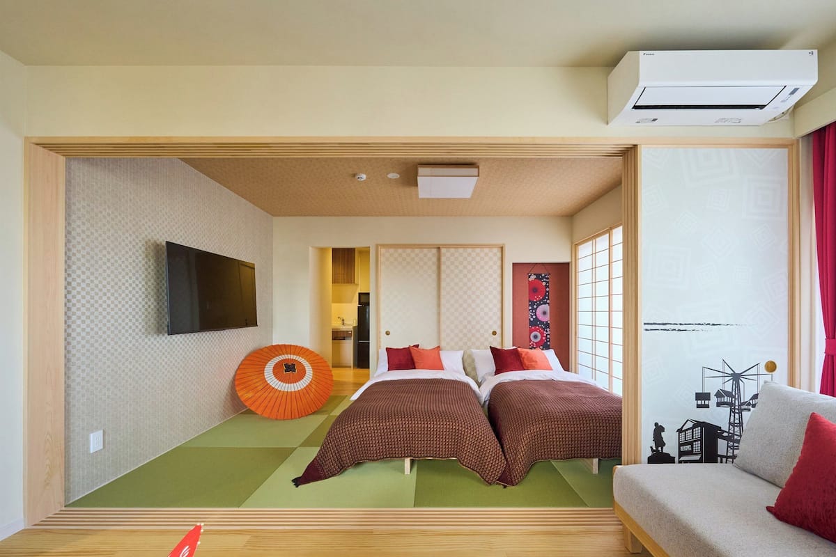 A hotel room with two beds, a sofa, a TV, an air conditioner, and a view of a traditional Japanese building.