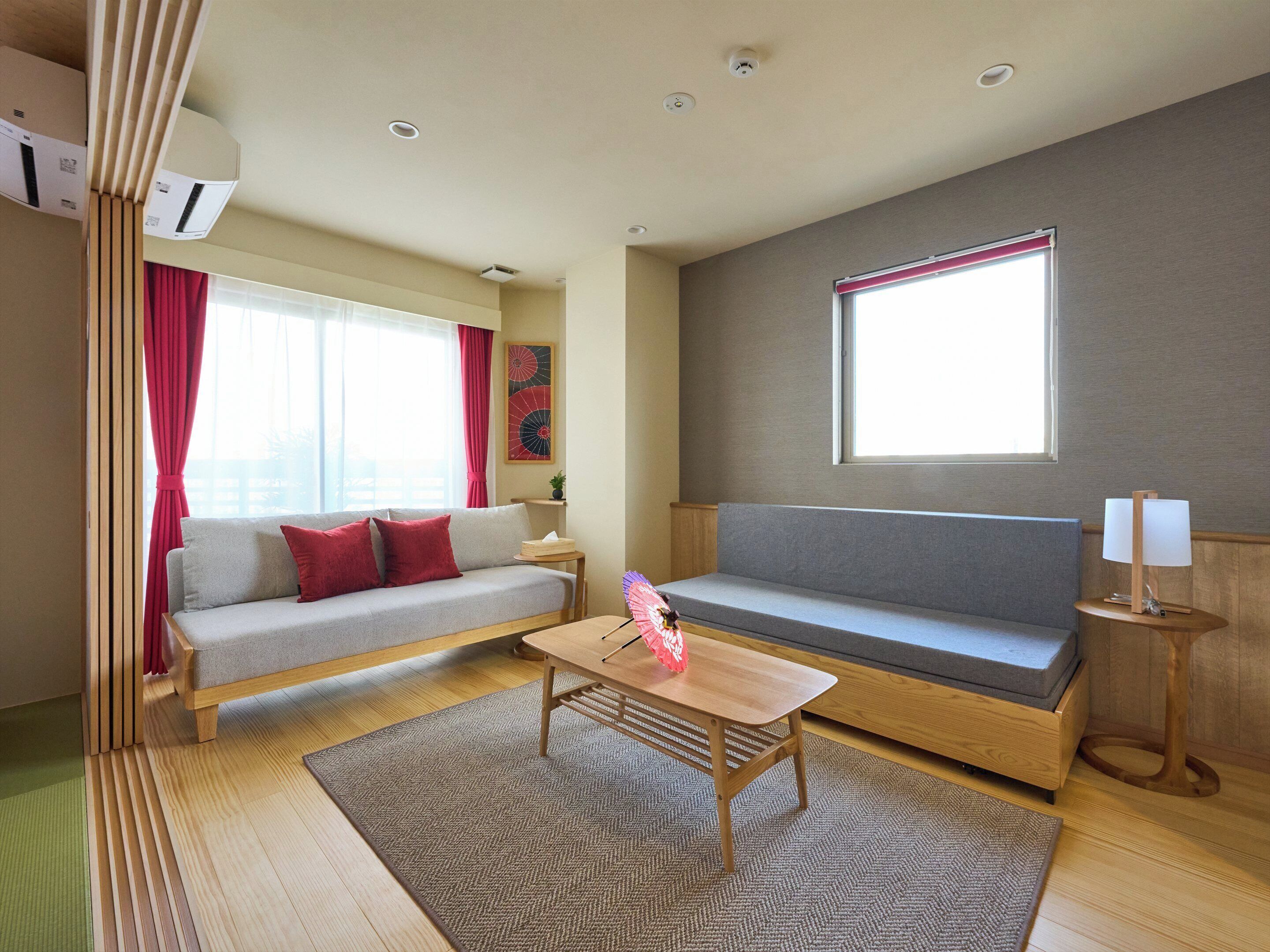 A modern living room with a grey sofa, a wooden coffee table, and a window with a red blind.