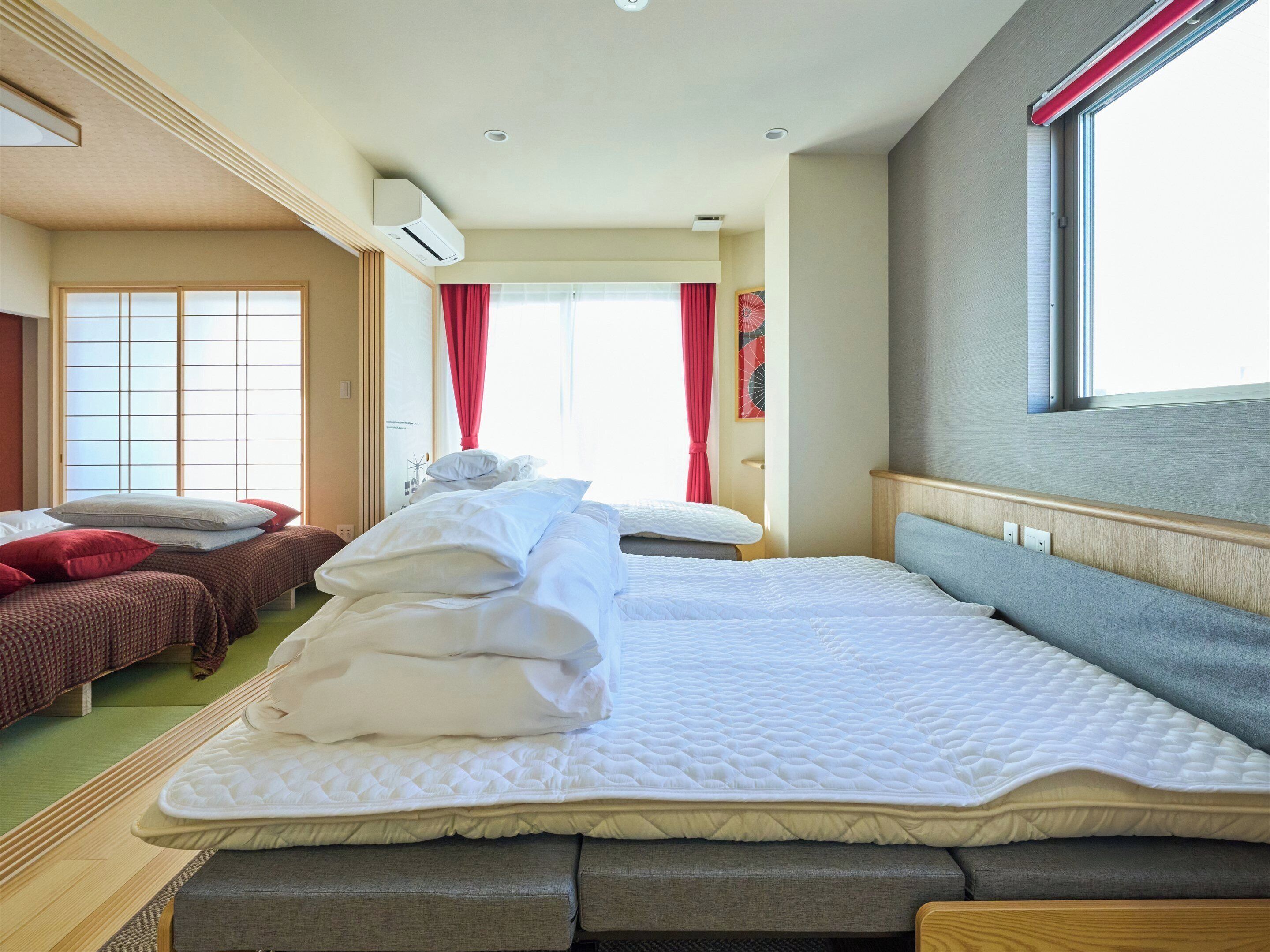 A hotel room with a large bed, a smaller bed, and a sofa bed. There is a window with curtains, an air conditioning unit, and a wall-mounted shelf.