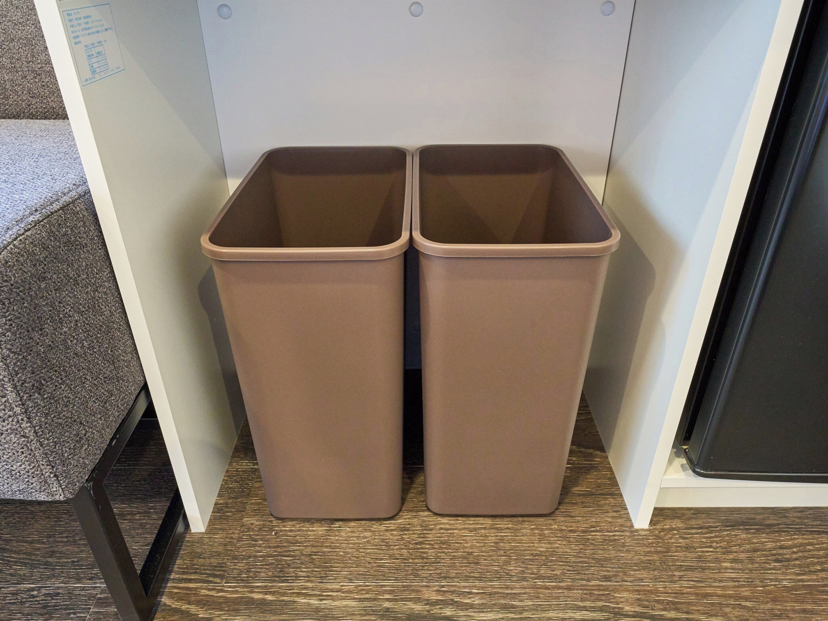 Two brown trash bins placed against a wall in a room with a wooden floor.