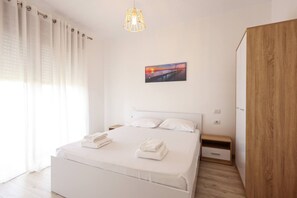Une chambre à coucher moderne avec un lit blanc, une armoire en bois, une table de chevet et un tableau représentant un coucher de soleil accroché au mur.