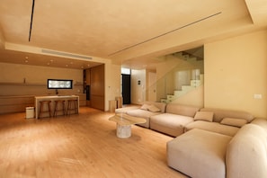 A modern living room with a wooden floor, a glass coffee table, a beige sofa, and a kitchen area with a TV and stools.
