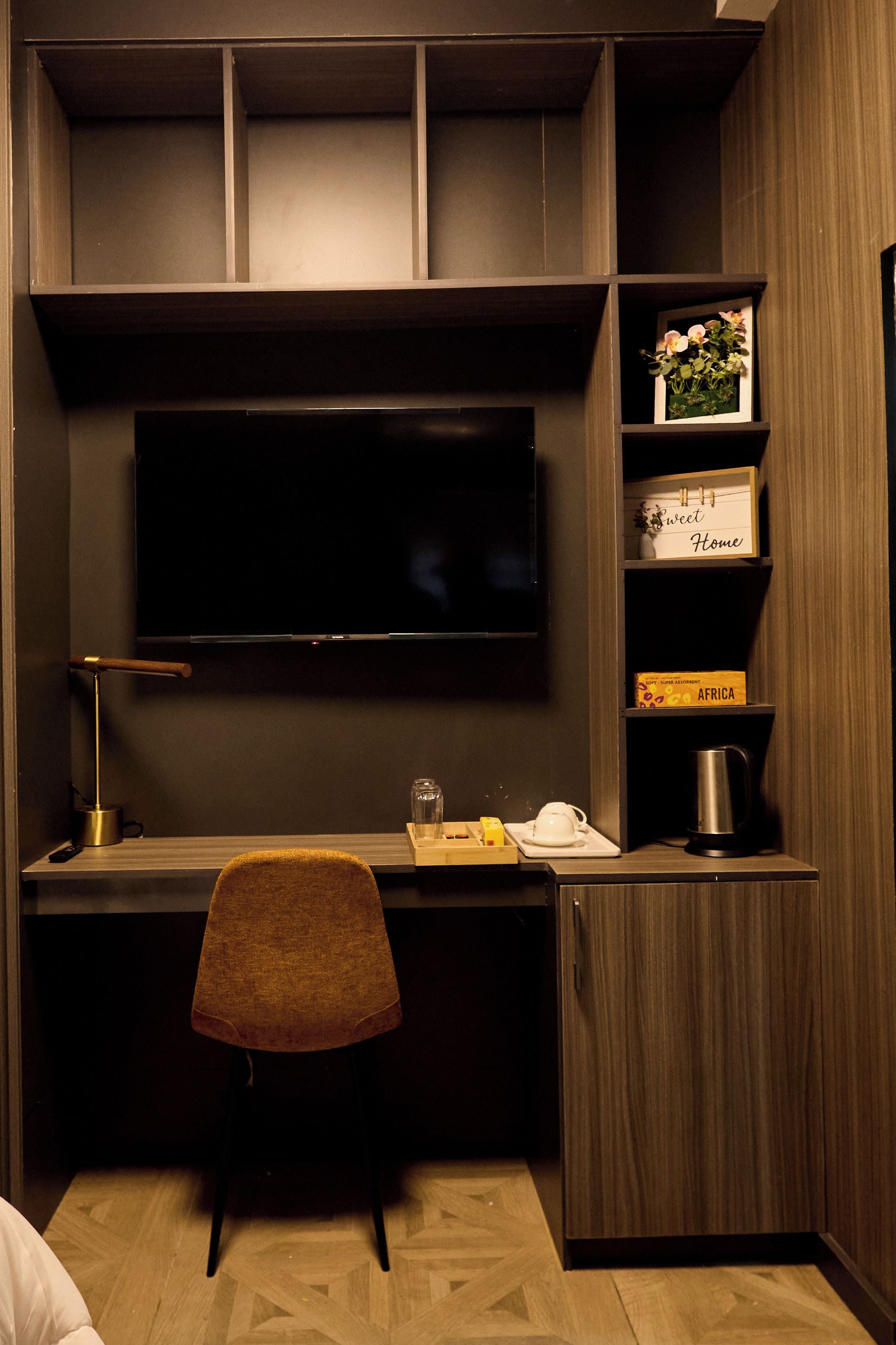 Une chambre moderne aux couleurs sombres, dotée d’un téléviseur à écran plat fixé au mur, d’un bureau avec une chaise, d’une machine à café et d’une étagère ornée d’objets décoratifs.