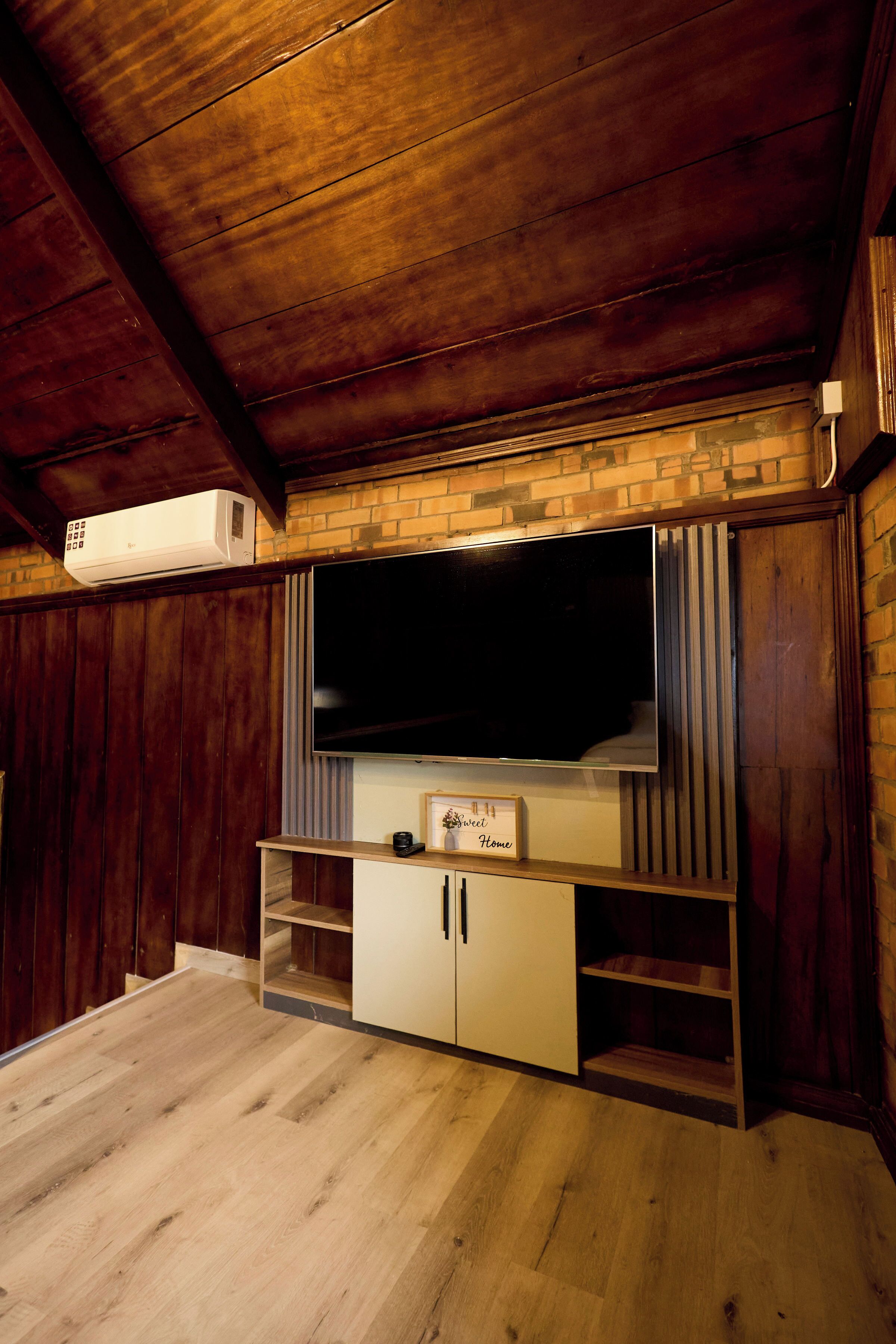 A room with a large flat-screen TV mounted on a brick wall, wooden flooring, and a built-in cabinet with a small shelf below the TV.