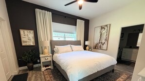 Een slaapkamer met een groot bed, een nachtkastje, een lamp, een kamerplant, een raam met gordijnen, een schilderij en een badkamer die door een geopende deur zichtbaar is.