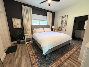 A bedroom with a large bed, a nightstand, a lamp, a potted plant, a window with curtains, a painting, and a bathroom visible through an open door.