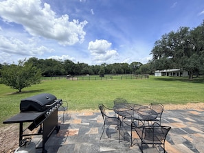 A patio with a grill and metal chairs, a white building with a porch, and a fenced pasture in the background.