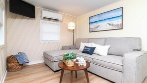 En stue med en beige sofa, et rundt træbord med en plante og pyngeting, et vægmonteret fjernsyn, en klimaanlæg og et maleri med et strandmotiv.