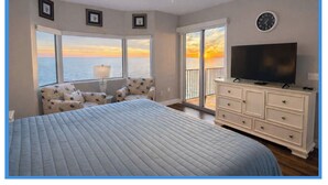 Une chambre avec un grand lit, une télévision, une commode et une vue sur l’océan au coucher du soleil.