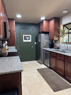 A kitchen with dark wooden cabinets, a stainless steel refrigerator, a granite countertop, and a tiled floor. - The Hidden Gem (Chico)