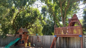 Un’area giochi in legno con scivolo e altalene, delimitata da una recinzione di legno e alberi.