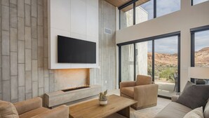 A modern living room with a flat-screen TV mounted on the wall, a fireplace, and large windows offering a view of the desert landscape. - Luxury Moab Casita with Resort Pool, Hot Tub & Bunk Room (Moab)