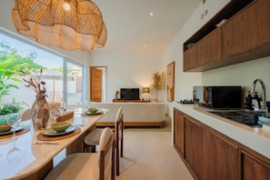 A modern kitchen with a dining area, wooden cabinets, a large window, and a television. - Lumi Villa by Betterplace (Pecatu)