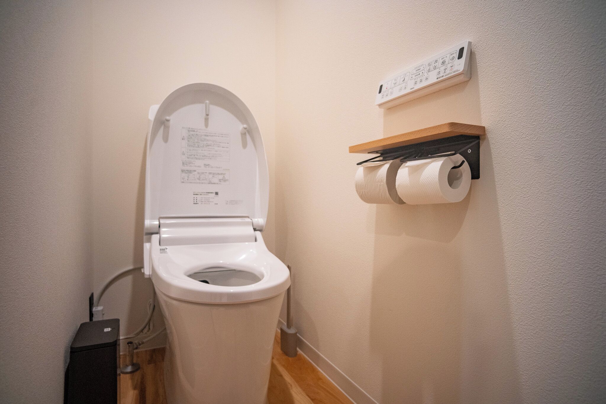 A toilet with a white lid, a wall-mounted paper towel dispenser, and a control panel above it.