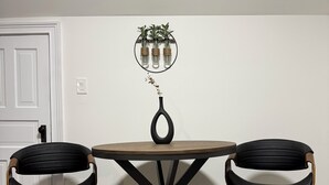 A dining table with a black metal frame and wooden legs, flanked by two black upholstered chairs with wooden armrests. Above the table, there is a wall-mounted mirror with a potted plant. - Spacious 6 bed 2 bath suburban escape 20 min from downtown Chicago (Blue Island)