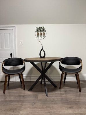 Una mesa de comedor con estructura metálica negra y patas de madera, flanqueada por dos sillas tapizadas negras con reposabrazos de madera. Sobre la mesa, un espejo montado en la pared con una planta en maceta.