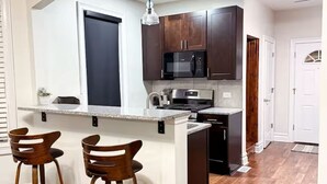 A kitchen with dark wooden cabinets, a granite countertop, and a built-in microwave. There are two bar stools and a hanging light fixture. - Spacious 6 bed 2 bath suburban escape 20 min from downtown Chicago (Blue Island)